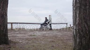 在户外轮椅上站在年轻男子旁边的白人女人的极长镜头。 残疾男孩和残疾儿童的侧视图